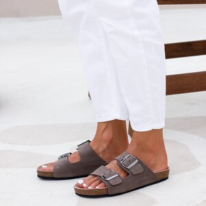 May include: A pair of brown leather sandals with two buckles on each strap. The sandals have a cork footbed and are worn on a person's feet.