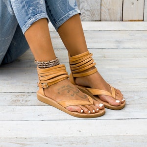 May include: Tan leather sandals with multiple straps around the ankle and a thong style toe strap. The sandals have a flat sole and are worn on a person's feet.