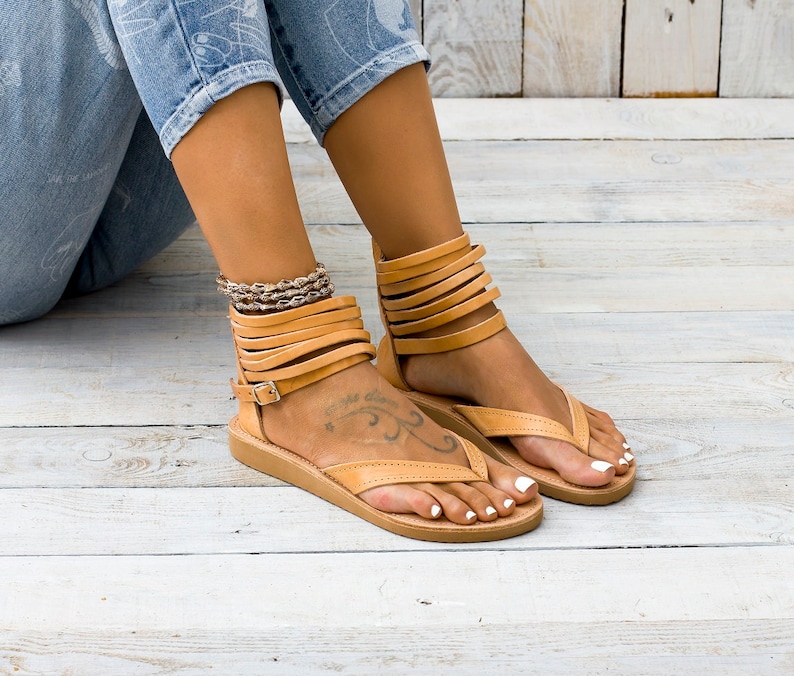 May include: Tan leather sandals with multiple straps around the ankle and a thong style toe strap. The sandals have a flat sole and are worn on a person's feet.
