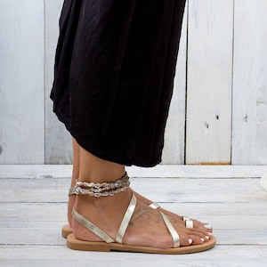 woman wears a two strap slip on pair of gold leather sandal with a toe ring. She is standing on a gray wooden floor.