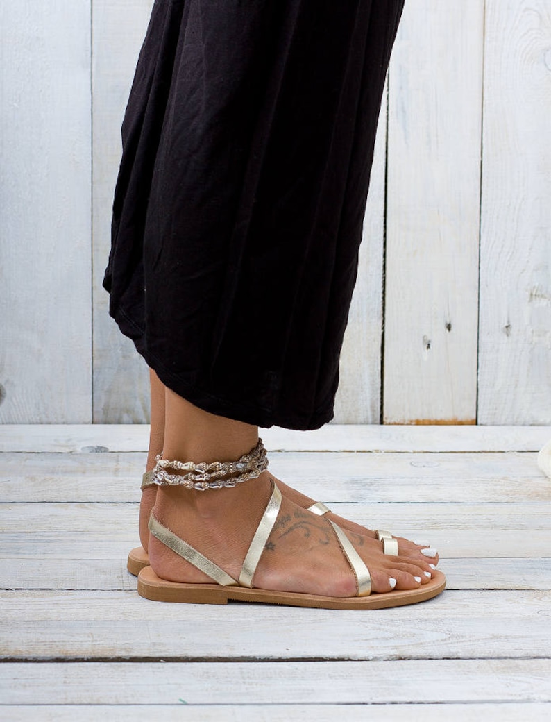 woman wears a two strap slip on pair of gold leather sandal with a toe ring. She is standing on a gray wooden floor.
