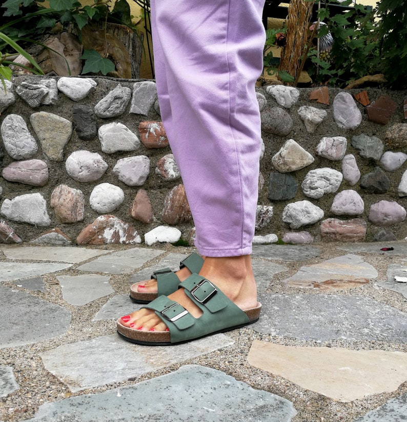 May include: A pair of green leather sandals with two buckles on each strap. The sandals are worn by a person wearing lavender pants.