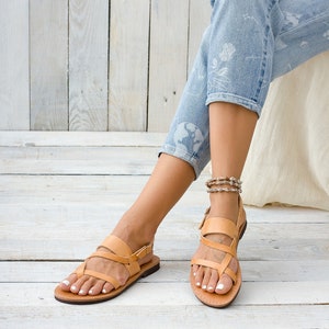 May include: Tan leather sandals with multiple straps and a buckle closure. The sandals are worn by a person with bare feet and are on a light wood floor.