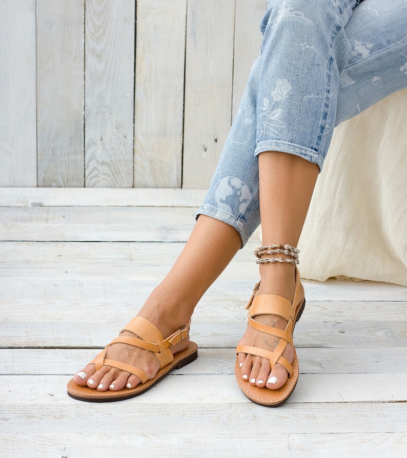 May include: Tan leather sandals with multiple straps and a buckle closure. The sandals are worn by a person with bare feet and are on a light wood floor.