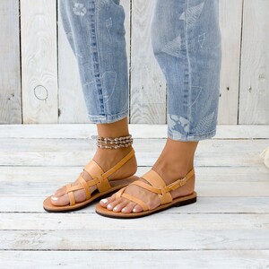 May include: Tan leather sandals with a criss-cross strap design and a buckle closure. The sandals are worn by a person with bare feet and are on a light wood floor.