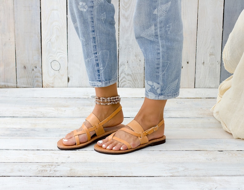 May include: Tan leather sandals with a criss-cross strap design and a buckle closure. The sandals are worn by a person with bare feet and are on a light wood floor.
