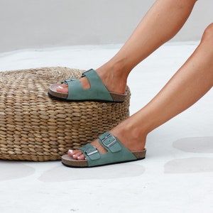 May include: A pair of sage green leather sandals with two adjustable buckles. The sandals have a cork footbed and a leather strap that goes around the ankle.