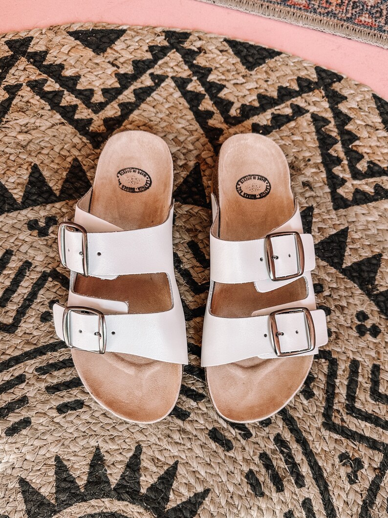 May include: A pair of white leather sandals with two adjustable buckles. The sandals have a cork footbed and are sitting on a woven rug.