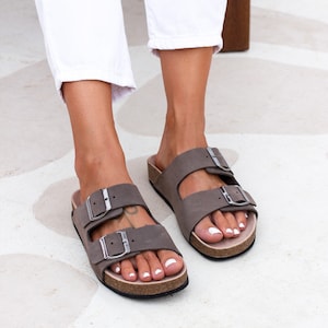 May include: A pair of brown leather sandals with two adjustable buckles. The sandals have a cork footbed and a white sole.