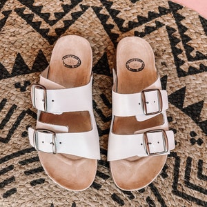 May include: A pair of white leather sandals with two adjustable buckles. The sandals have a cork footbed and are sitting on a woven rug.