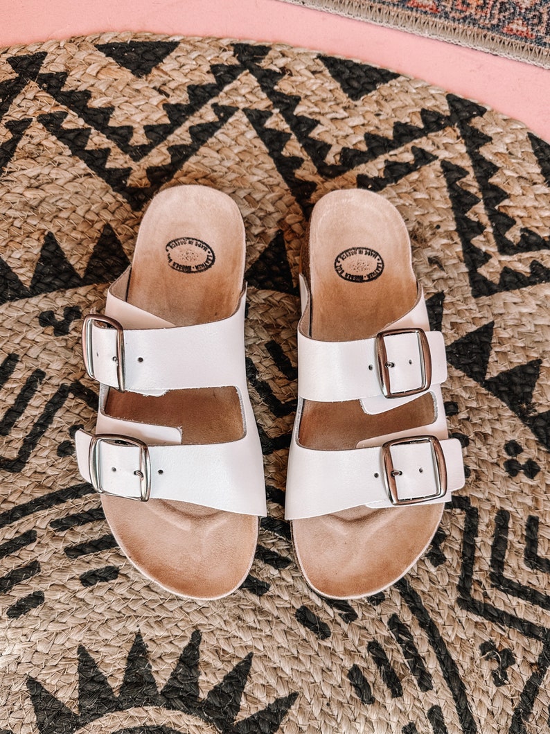 May include: A pair of white leather sandals with two adjustable buckles. The sandals have a cork footbed and are sitting on a woven rug.