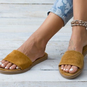May include: A pair of brown suede slide sandals with a flat sole. The sandals are worn by a person with a shell anklet.