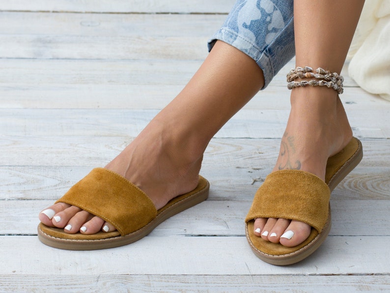 May include: A pair of brown suede slide sandals with a flat sole. The sandals are worn by a person with a shell anklet.