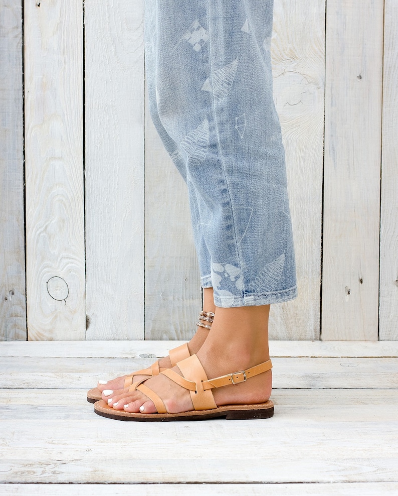 May include: Tan leather sandals with multiple straps and a buckle closure. The sandals are worn by a person wearing light blue jeans with a white pattern.