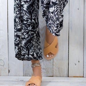 May include: A pair of tan leather sandals with a criss-cross strap design. The sandals are being worn by someone wearing black and white floral trousers.