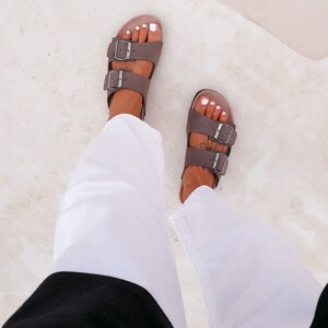 May include: A pair of brown leather sandals with two buckles on each strap. The sandals are worn by a person wearing white pants and a black top.