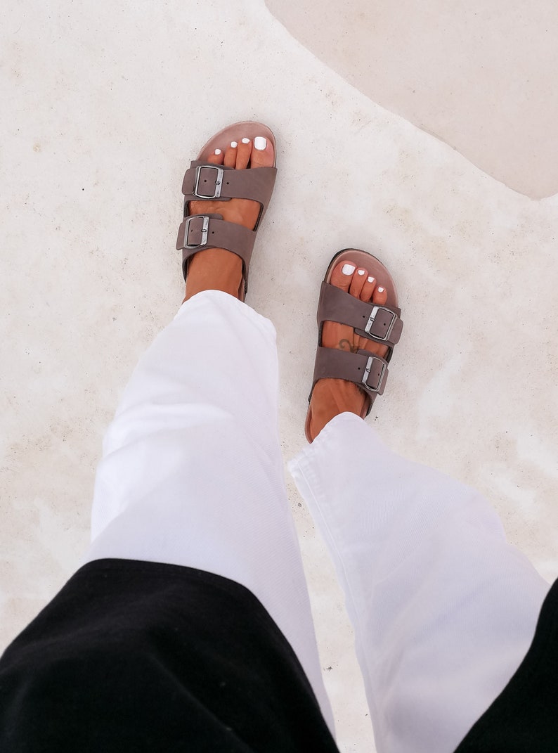 May include: A pair of brown leather sandals with two buckles on each strap. The sandals are worn by a person wearing white pants and a black top.