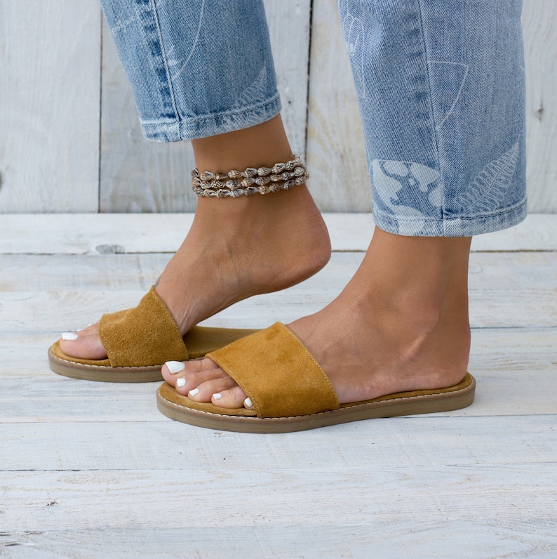 May include: A pair of brown suede slide sandals with a flat sole. The sandals are worn by a person with bare feet and a shell anklet.