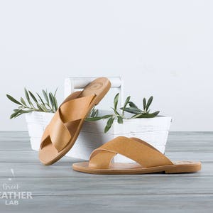 May include: Two pairs of tan leather sandals with criss-cross straps. The sandals are sitting on a white wooden shelf with green leaves.