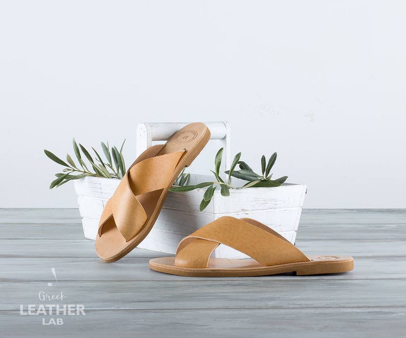 May include: Two pairs of tan leather sandals with criss-cross straps. The sandals are sitting on a white wooden shelf with green leaves.
