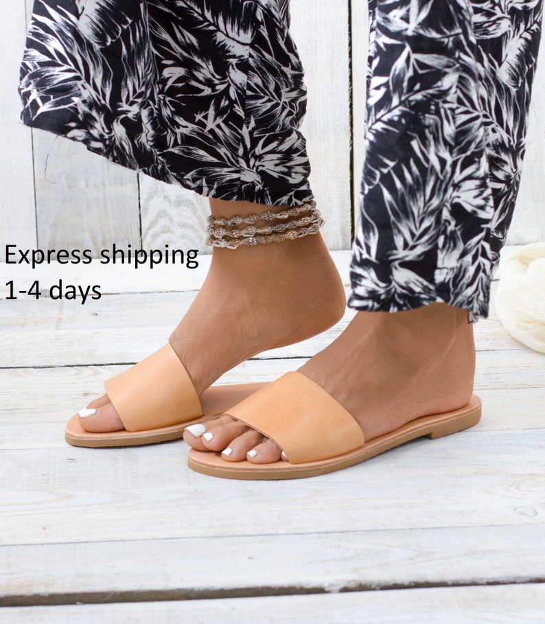 May include: A pair of light brown leather sandals with a simple design. The sandals have a flat sole and a single strap across the top of the foot.