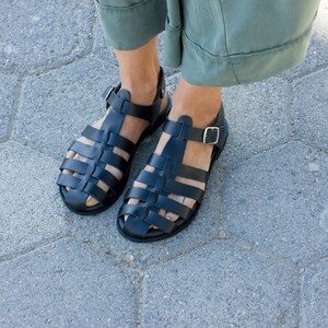 Puede incluir: Un par de sandalias gladiadoras de cuero negro con cierre de hebilla. Las sandalias tienen un dise&ntilde;o tejido y se llevan en los pies.
