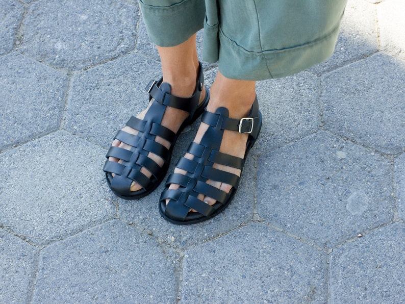 Puede incluir: Un par de sandalias gladiadoras de cuero negro con cierre de hebilla. Las sandalias tienen un dise&ntilde;o tejido y se llevan en los pies.