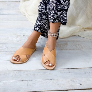May include: A pair of tan leather sandals with criss-cross straps. The sandals are being worn by a person with bare feet and white nail polish.
