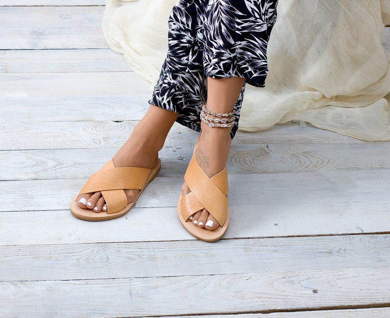 May include: A pair of tan leather sandals with criss-cross straps. The sandals are being worn by a person with bare feet and white nail polish.