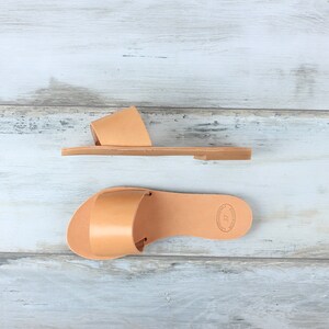 May include: Two pairs of tan leather sandals with a simple design. The sandals have a single strap across the top of the foot and a flat sole.