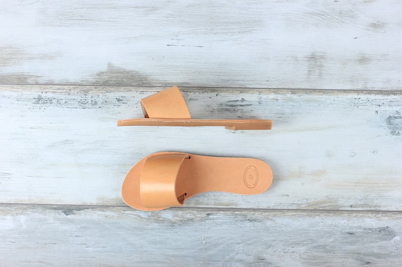 May include: Two pairs of tan leather sandals with a simple design. The sandals have a single strap across the top of the foot and a flat sole.
