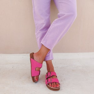 May include: A pair of pink leather sandals with two straps and buckles. The sandals are worn by a person wearing lavender pants.