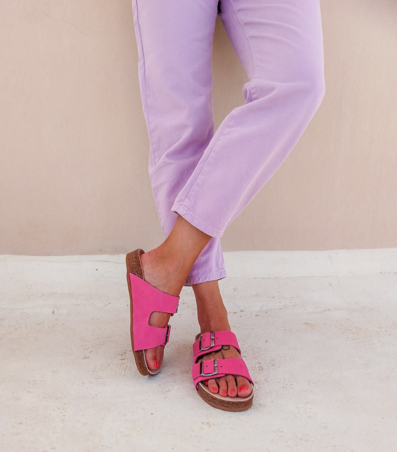 May include: A pair of pink leather sandals with two straps and buckles. The sandals are worn by a person wearing lavender pants.