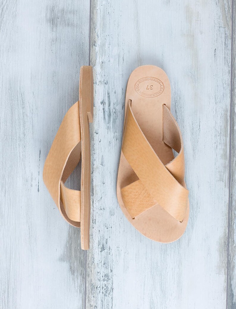 May include: A pair of tan leather sandals with a criss-cross strap design. The sandals have a simple, minimalist style and are made of high-quality leather.