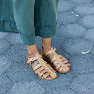 Puede incluir: Un par de sandalias de cuero marr&oacute;n con un dise&ntilde;o enjaulado y cierres de hebilla. Las sandalias son usadas por una persona de pie sobre una superficie de baldosas grises.