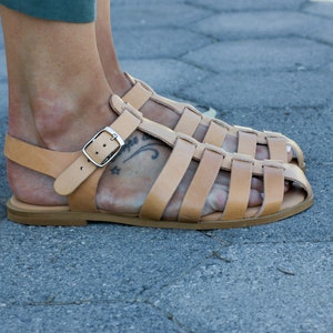 Puede incluir: Sandalias gladiadoras de cuero color marr&oacute;n claro con cierre de hebilla. Las sandalias tienen una suela gruesa y se llevan en los pies.