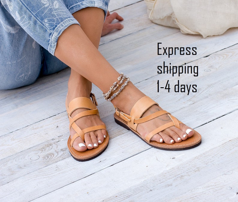 May include: Tan leather sandals with a toe thong and multiple straps. The sandals are worn on a pair of feet with white nail polish. The sandals are on a light wood floor.