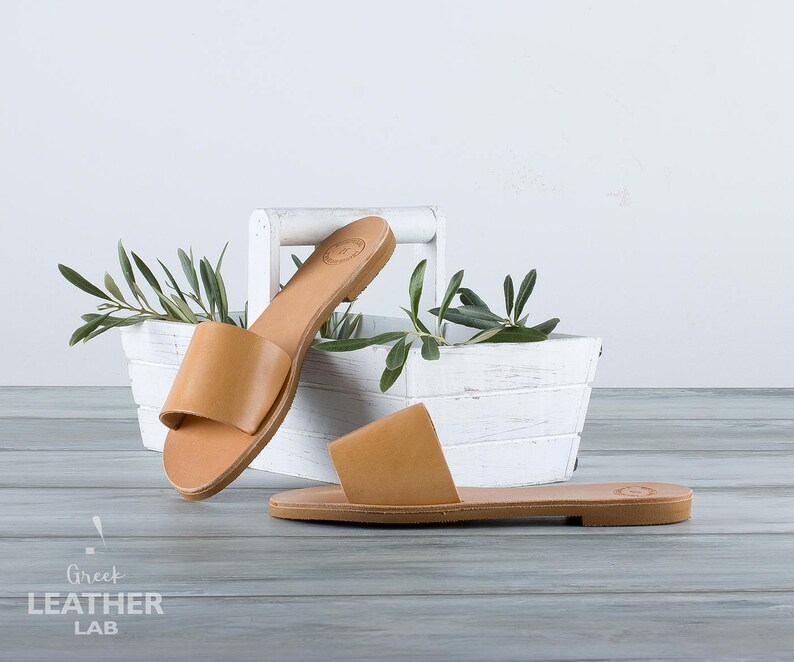 May include: Two pairs of tan leather sandals with a simple design. The sandals are on a wooden surface with a white planter with green plants in the background.