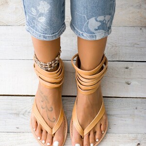 May include: Tan leather sandals with multiple straps around the ankle. The sandals have a thong style toe strap and are worn on a pair of feet with white nail polish.