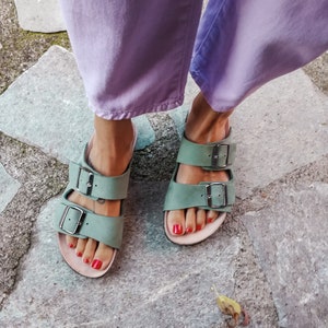 May include: A pair of green leather sandals with two buckles on each strap. The sandals are worn by a person wearing lavender pants.