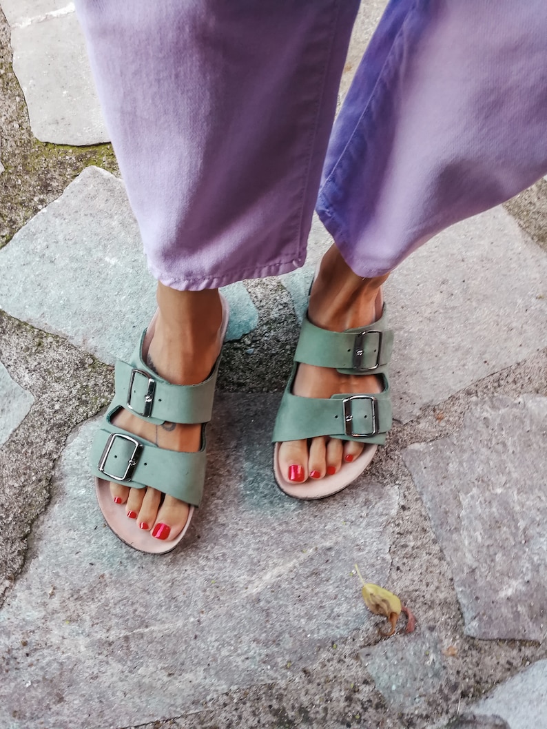 May include: A pair of green leather sandals with two buckles on each strap. The sandals are worn by a person wearing lavender pants.