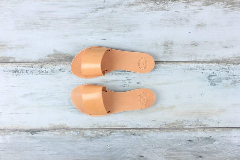 May include: A pair of tan leather sandals with a simple design. The sandals have a flat sole and a strap across the top of the foot.