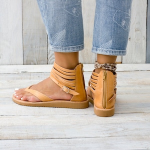 May include: Tan leather sandals with multiple straps and a zipper on the back. The sandals have a flat sole and are worn by a person with denim jeans.