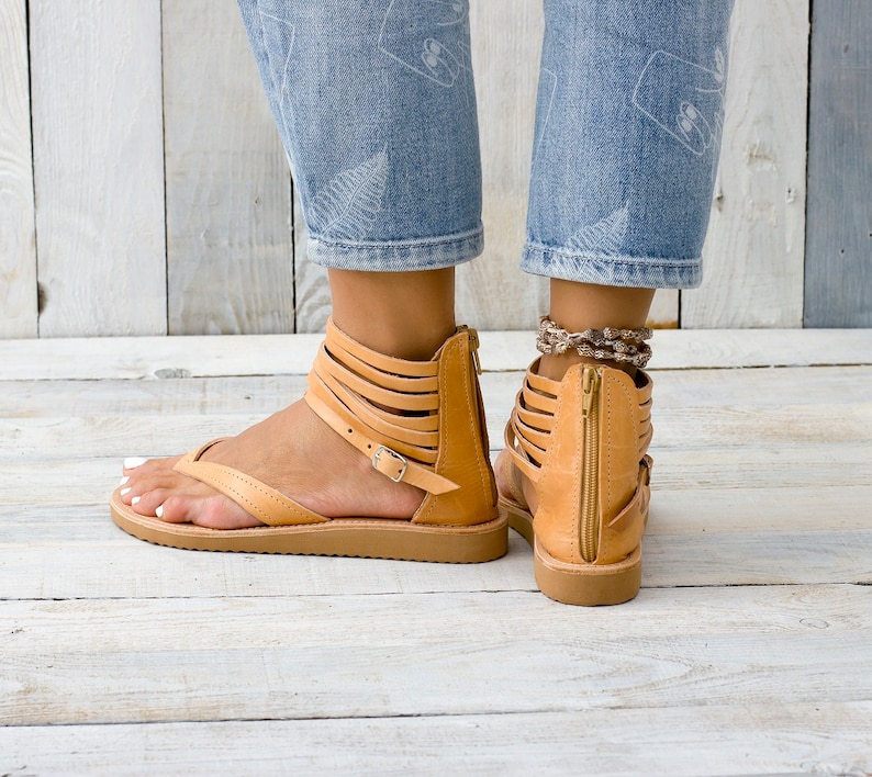 May include: Tan leather sandals with multiple straps and a zipper on the back. The sandals have a flat sole and are worn by a person with denim jeans.
