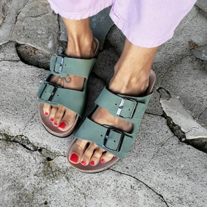 May include: A pair of green leather sandals with two adjustable buckles. The sandals have a cork footbed and are worn on a person's feet with red nail polish.