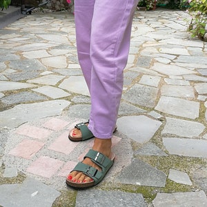 May include: A pair of green leather sandals with two buckles on each strap. The sandals are worn by a person wearing lavender pants.