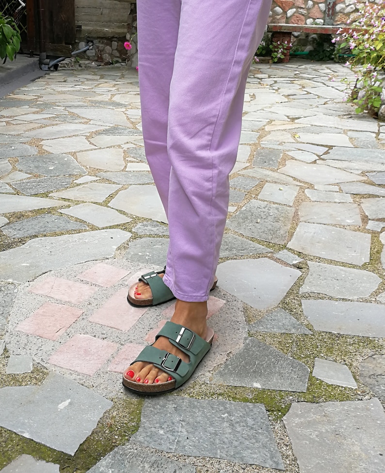 May include: A pair of green leather sandals with two buckles on each strap. The sandals are worn by a person wearing lavender pants.