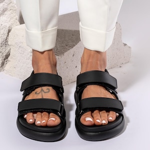 May include: Black sandals with two adjustable straps. The sandals have a thick sole and are worn with white pants. The image is a close-up of the feet and sandals.