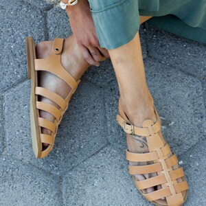 Puede incluir: Un par de sandalias de cuero marr&oacute;n con un dise&ntilde;o enjaulado y cierres de hebilla. Las sandalias se llevan en los pies de una persona, con un pie apoyado en una superficie de hormig&oacute;n gris.