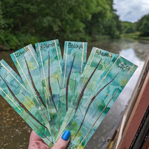 Könnte beinhalten: Ein Set aus sechs Aquarell-Lesezeichen mit einem grünen und blauen Aquarell-Design mit goldenen Akzenten. Die Lesezeichen sind in einer Hand aufgefächert.